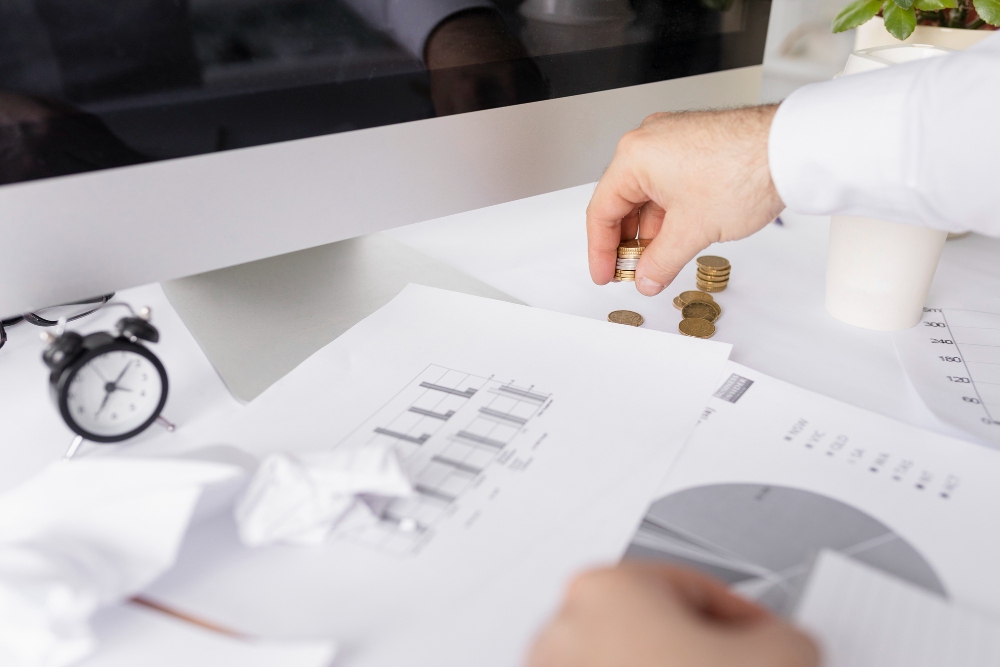 Mężczyzna liczący monety przy biurku z dokumentami finansowymi, wykreślnikami i monitorem komputera